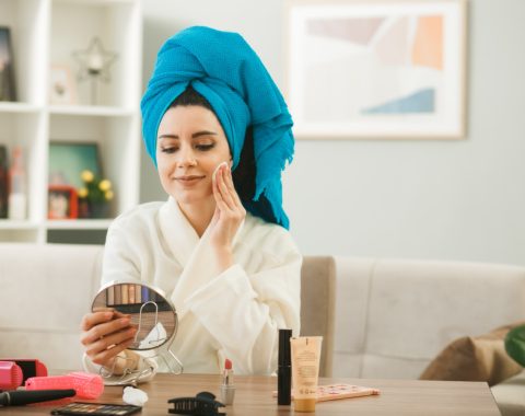 pleased-young-girl-holding-looking-mirror-applying-tone-up-cream-with-sponge-sitting-table-with-makeup-tools-living-room (1)